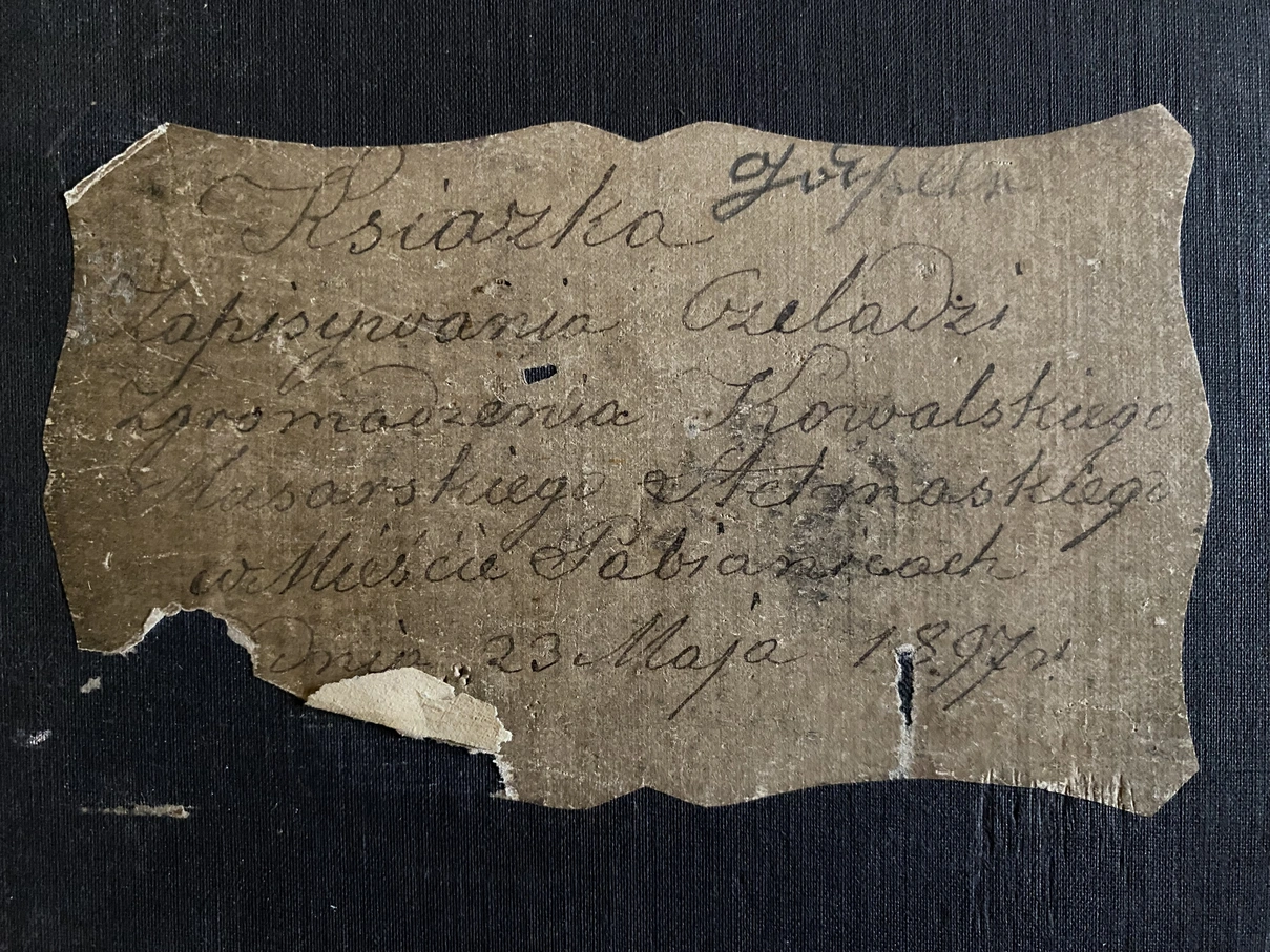 Fragment czarnej oprawy z etykietą. Na etykiecie tytuł „Książka zapisywania czeladzi Zgromadzenia Kowalskiego Ślusarskiego Stelmaskiego w mieście Pabianicach” oraz data 1897